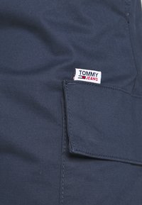 Tmavě modré kalhoty s přední kapsou, na které je bílá etiketa s nápisem "TOMMY JEANS" červeně a modře. Hladká textura látky.