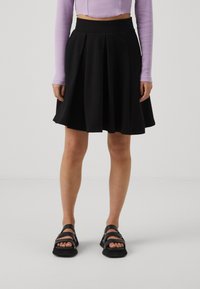 Black pleated skirt with a fitted waistband, made from smooth material; paired with chunky black sandals and a purple long-sleeve top.