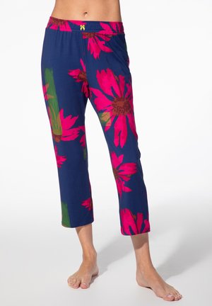 Blaue gekürzte Hosen mit großen, leuchtend pinken und grünen Blumenmustern, getragen von einer barfuß laufenden Person.