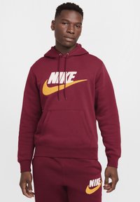 Sudadera con capucha burdeos con un gran logo de Nike en blanco y amarillo, bolsillo canguro, capucha con cordón y puños acanalados. Textura de tela suave.
