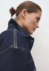 Chaqueta de mezclilla en azul oscuro con costuras amarillas en contraste; cuenta con un cuello, cierre de botones a presión y forma clásica.