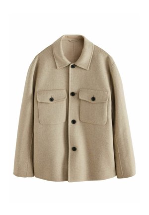 Mand iført beige uldjakke med knapper, lyserød skjorte, mørkegrå bukser og sorte læder loafers, stående udendørs ved en stenvæg.