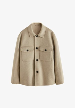 Homme portant une veste en laine beige boutonnée, une chemise rose clair, un pantalon gris foncé et des mocassins en cuir noir, debout dehors près d'un mur en pierre.