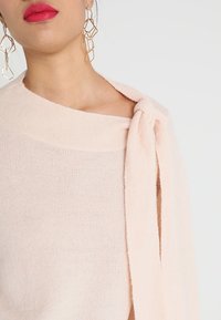 Weicher rosa gestrickter Pullover mit asymmetrischem Design, der ein Bindeband an einer Schulter und einen weiten Halsausschnitt aufweist. Strukturiert sichtbares Material.