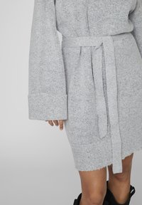 Robe en tricot gris clair à manches longues avec une ceinture nouée à la taille, visible de la mi-cuisse à la main, avec une partie des bottes noires visibles.
