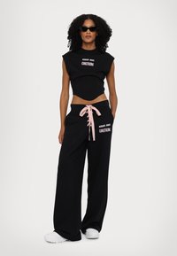 Zwarte cropped top met reliëf tekst en zwarte sweatpants met wijde pijpen, uitgerust met een roze trekkoord en een logo op het linkerpijp. Witte sneakers maken de look af.