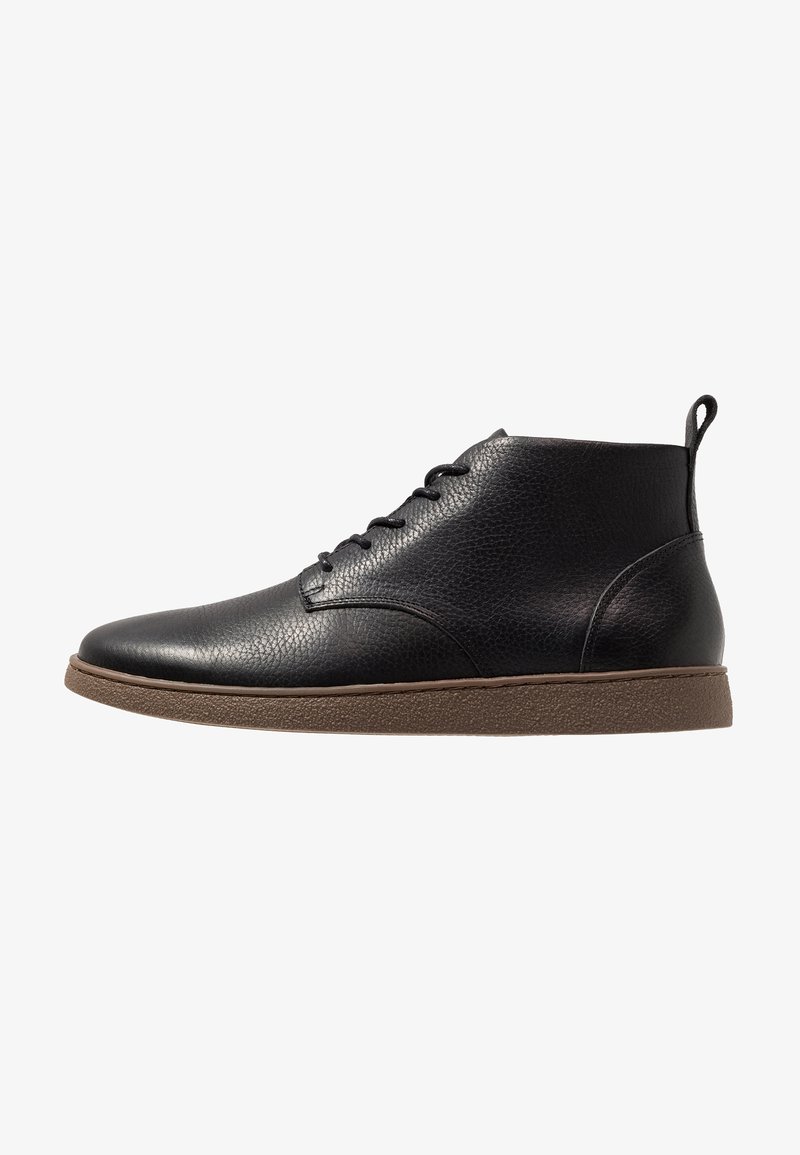 Bottine noire en cuir avec semelle en caoutchouc marron, laçage à l'avant, surface texturée et languette au talon sur fond blanc.