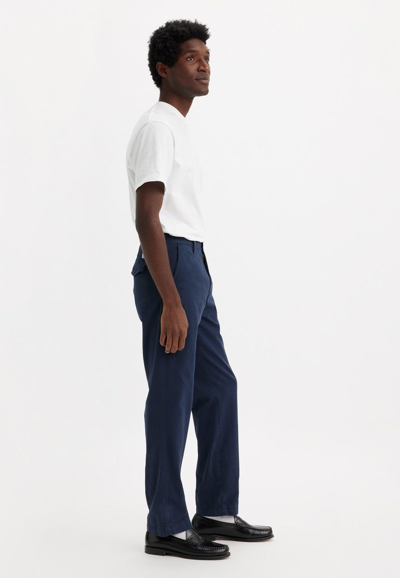 Camiseta blanca, pantalones azul marino y mocasines negros con calcetines blancos. Los pantalones tienen un corte recto y una textura sutil, con bolsillos laterales.