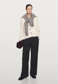 Crème cardigan met donkere knopen, grijze sjaal over de schouders gedrapeerd, wijde zwarte broek en een donkere clutch. Zachte texturen overal.