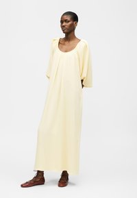 Γυναίκα που φοράει ένα χαλαρό, ανοιχτό κίτρινο μακρύ φόρεμα με φουσκωτά μανίκια και καφέ δερμάτινα παπούτσια με κορδόνια, σε λευκό φόντο.