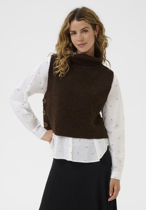 Brown, ribbed knit sleeveless sweater with a high collar and three large buttons on the side, worn over a white, patterned long-sleeve shirt.