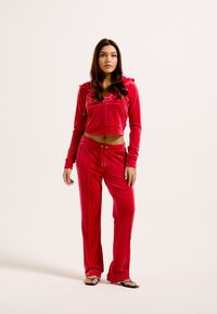Tuta abbinato in velluto rosso, composto da un hoodie cropped con zip e pantaloni con elastico in vita e lacci. Il tessuto ha una texture liscia.