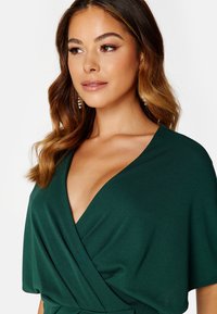 Groene wikkeltop met een diepe V-hals en korte mouwen, gemaakt van een zachte stof met een gladde textuur en subtiele draperingdetails.