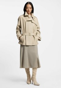 Gabardina beige con cinturón, grandes solapas y un bolsillo parche, combinada con una falda gris asimétrica y botas de tacón alto hasta la rodilla en color crema.