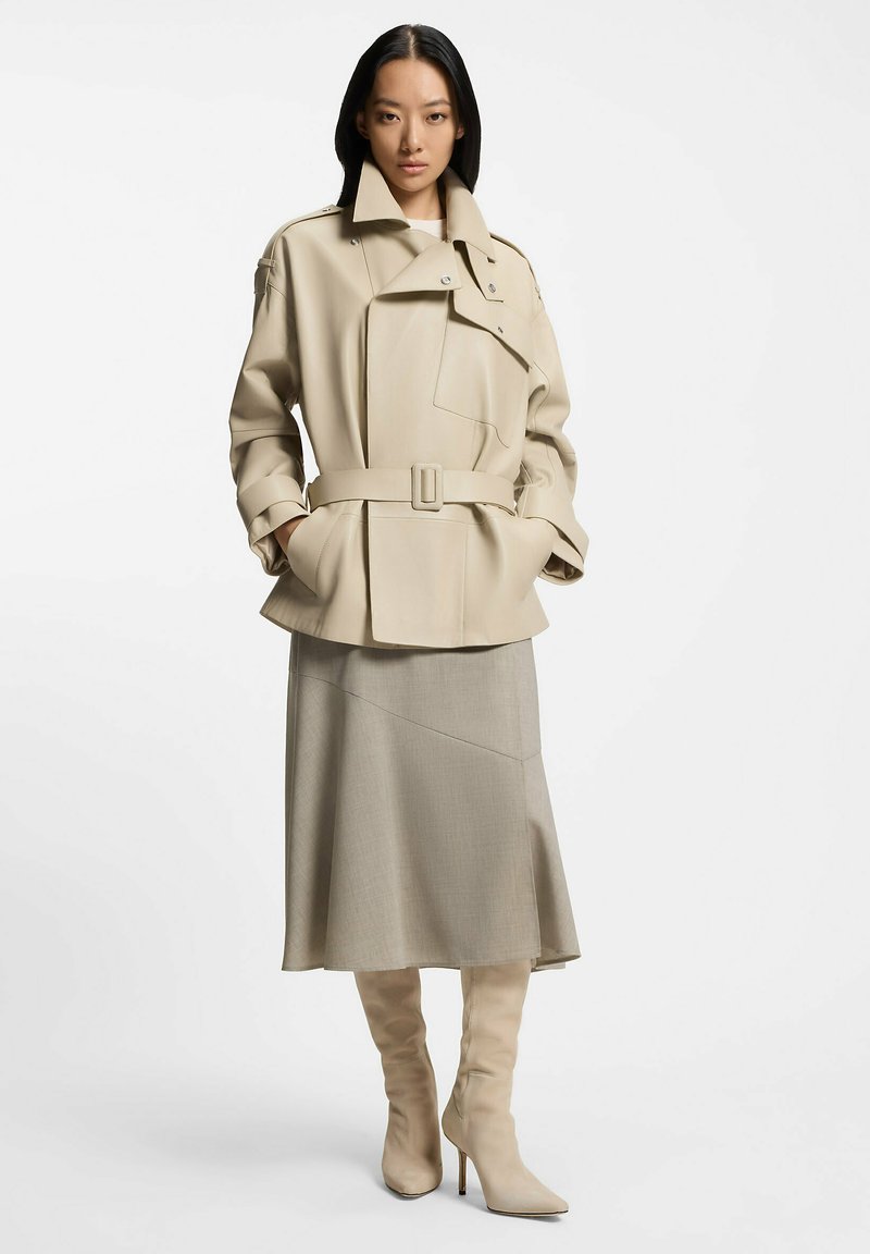 Gabardina beige con cinturón, grandes solapas y un bolsillo parche, combinada con una falda gris asimétrica y botas de tacón alto hasta la rodilla en color crema.