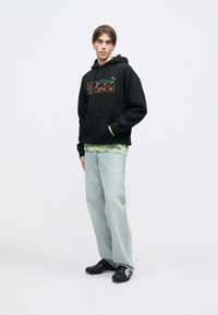 Melna kapuce ar krāsainu grafisko dizainu, virsū uz gaiši zaļas krāsas tie-dye krekla. Gaiši zili džinsi un melna apģērba apavi pabeidz šo tērpu.