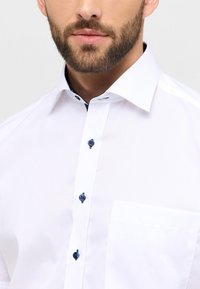 Camicia bianca con colletto alla francese, dotata di bottoni blu scuro e un accento testurizzato all'interno del colletto. Include una tasca sul petto.