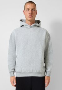 Uomo che indossa un hoodie grigio chiaro con tasca frontale e pantaloni neri, in piedi contro uno sfondo bianco semplice.