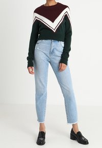 Persona che indossa un pullover a zig zag bordeaux, bianco e verde, jeans chiari a vita alta e mocassini neri con tacco e dettagli decorativi.