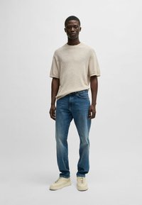 Beige ribbed short-sleeve sweater paired with light blue denim jeans. Model wears beige sneakers with a minimal design. Simple background.