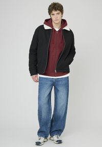 Veste noire avec un col en fourrure blanche portée sur un hoodie bordeaux, un t-shirt blanc et un jean bleu large. Chaussures dans un mélange de blanc et de bleu marine.