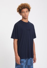 Jeune homme aux cheveux bouclés portant un t-shirt ample bleu marine foncé et un pantalon bleu à motifs sur un fond clair uni.