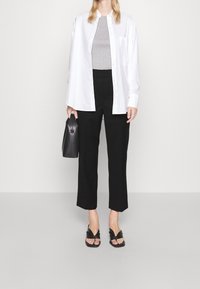 Femme portant une chemise blanche à boutons sur un haut gris, un pantalon noir coupé, des talons ouverts noirs, tenant un sac à main noir, debout sur un sol gris.