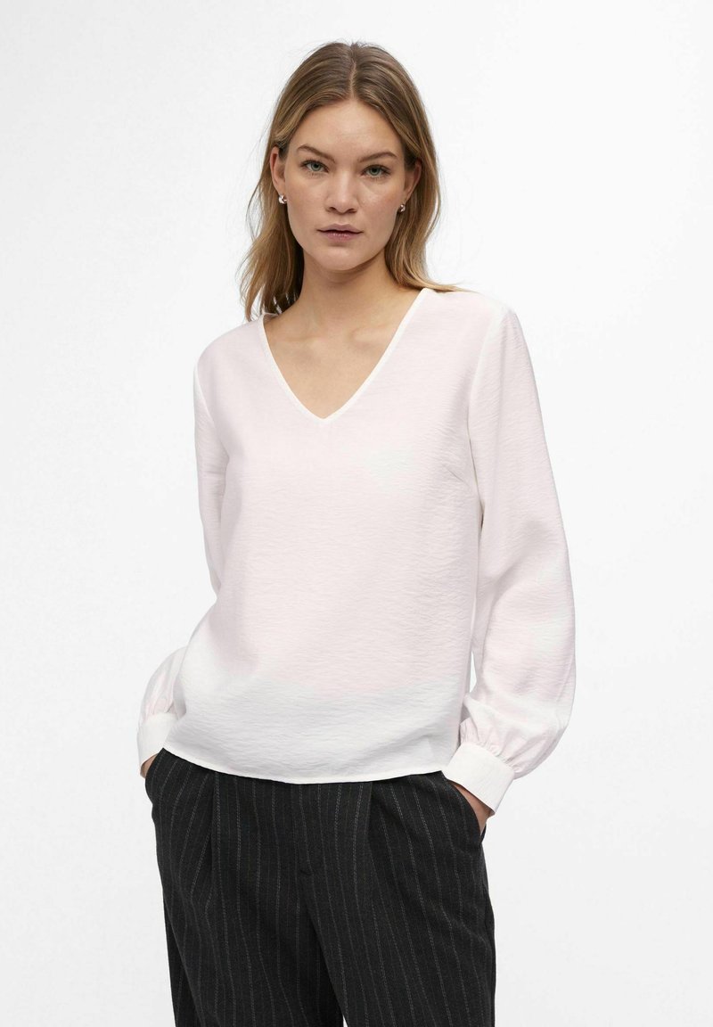 Lys pink v-neck bluse med lange ærmer og tekstureret stof, der har hvide manchetter. Parret med sorte bukser med striber.