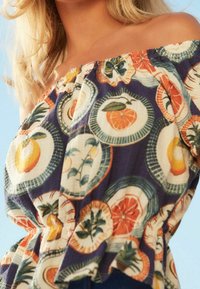 Off-shoulder top med et farverigt frugtmønster på en mørk baggrund, lavet af et let materiale med en tekstureret overflade.