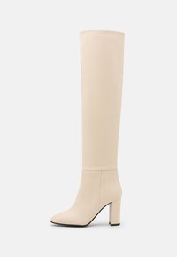 Botas beige por encima de la rodilla hechas de cuero suave, con punta afilada y un tacón alto y grueso. Diseño minimalista sin acentos visibles.