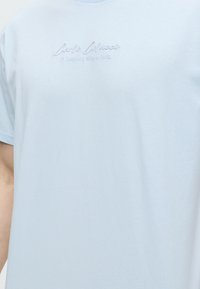 Ανοιχτό μπλε μπλουζάκι με κεντημένο κείμενο "Carlo Colucci A Legacy Since 1978" στο στήθος.