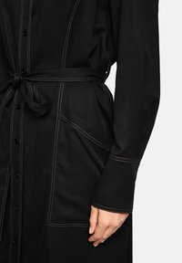 Schwarzes Hemdkleid mit Bindegürtel, kontrastierenden Nähten, Knopfverschluss und zwei großen Fronttaschen auf beiden Seiten.