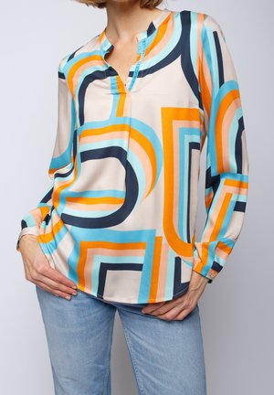 Mujer que lleva una blusa de manga larga con un patrón geométrico colorido en naranja, azul, negro y beige combinada con pantalones vaqueros azul claro.