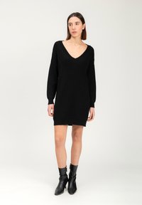 Abito nero lavorato a maglia con maniche lunghe e scollatura a V profonda. Caratterizzato da motivi testurizzati e una vestibilità comoda, arriva a metà coscia. Indossato con stivali neri alla caviglia.