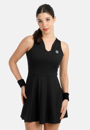 Donna con capelli scuri raccolti in una coda di cavallo, indossa un vestito da tennis nero senza maniche e polsini neri, in piedi contro uno sfondo chiaro e uniforme.