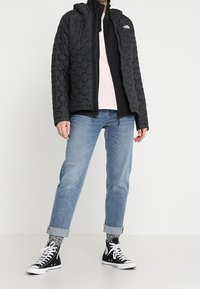 Giacca nera trapuntata con cappuccio, abbinata a una camicia rosa chiaro, jeans blu e sneakers alte nere. Outfit casual a strati.