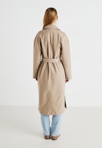 Abrigo de lana beige hasta la rodilla con cinturón en la cintura, cuello ancho y aberturas laterales; combinado con vaqueros azul claro y vista posterior visible.