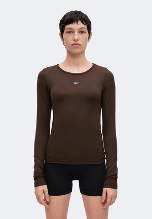Junge Person mit kurzen dunklen Haaren, trägt ein eng anliegendes braunes, geripptes Langarmshirt und schwarze Shorts, steht vor einem einfarbigen hellen Hintergrund.