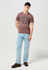 Kortärmad brun T-shirt med svart "WRANGLER 1947" grafik, bärs med ljusblå jeans och bruna skor. Slät textur. Avslappnad passform.