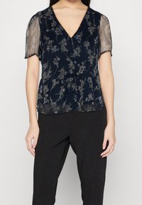 Blusa floreale navy con maniche corte trasparenti, scollatura a V e vita arricciata. Presenta motivi floreali sottili in colori chiari. Abbinata a pantaloni neri.