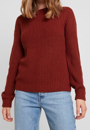 Frau trägt einen strukturierten, rostfarbenen Strickpullover und hellblaue Jeans und steht vor einem einfarbigen hellgrauen Hintergrund.