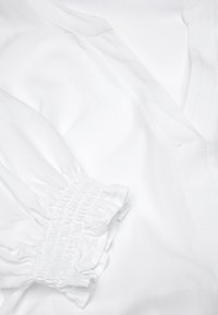 Blusa bianca con scollo a V e maniche arricciate. Tessuto liscio con una leggera lucentezza e dettagli di arricciatura ai polsini.