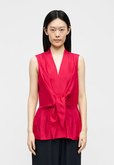 Femme aux cheveux noirs raides porte un haut rouge sans manches avec un nœud à l'avant et un pantalon noir, debout devant un fond clair uni.