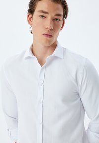 Camisa blanca de botón con una tela texturizada, mangas largas arremangadas en los puños y un cuello clásico. Presenta un cierre frontal con botones.