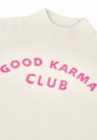 Crèmekleurige trui met een ribbelige opstaande kraag en roze fuzzy letters, met de tekst "GOOD KARMA CLUB" in een gebogen ontwerp.