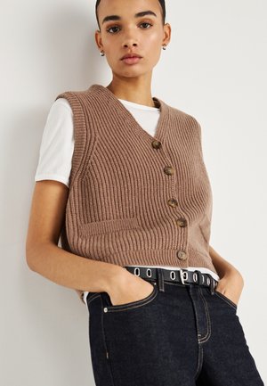 Rjava rebrasta pletena telovnik z V-izrezom, petimi gumbi in stranskimi žepi, nošen čez belo srajco, kombiniran s temnimi denim jeans hlačami in črnim pasom.