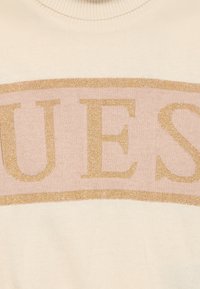 Maglione crema con scollatura rotonda caratterizzato da una fascia prominente rosa chiaro. La parola "GUESS" è mostrata in glitter dorato. Texture morbida.