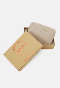 Valentino beige zipplåda i en brun pappask. Utmärks av guldbranding och slät textur. Asken märkt med återvinningsanvisningar.