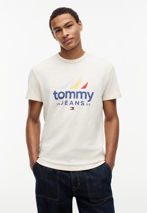 Młody mężczyzna w jasnobeżowej koszulce Tommy Jeans z wielokolorową grafiką i ciemnoniebieskich dżinsach, ręce w kieszeniach, neutralne tło.