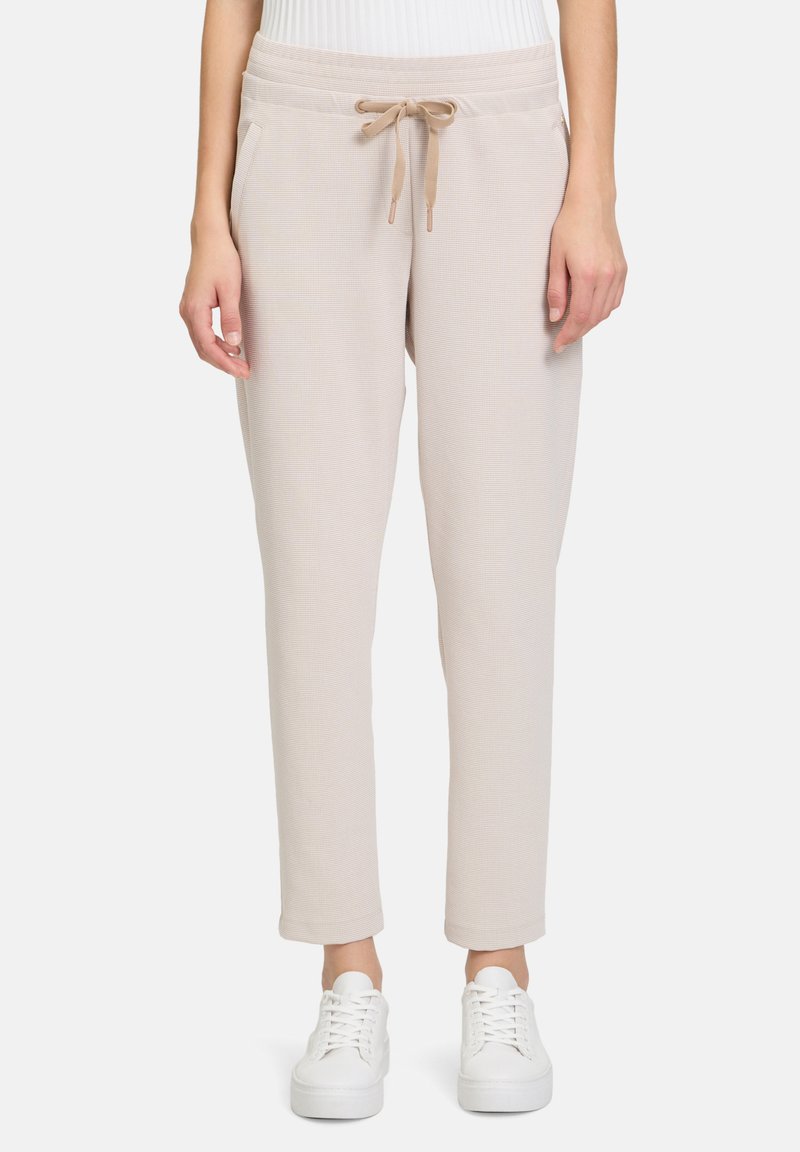 Beige gewebte Jogginghosen mit elastischem Taillenbund, Kordelzugdetails, Seitentaschen und schmal zulaufenden Bündchen, kombiniert mit weißen Sneakers.